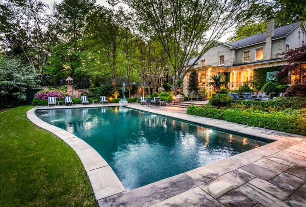 A luxurious backyard with a rectangular swimming pool, surrounded by lush greenery, patio chairs, and a large house in the background