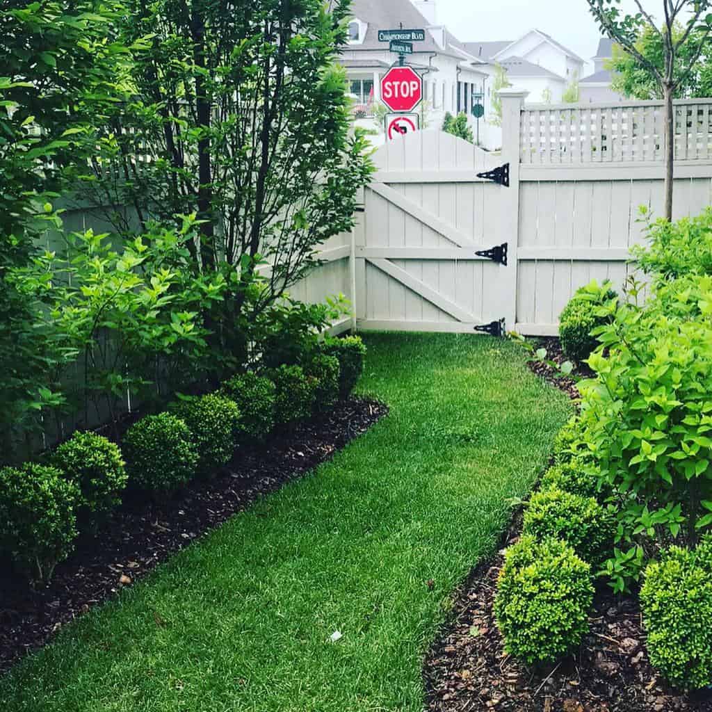 Side yard with a lush green lawn, neatly trimmed shrubs, a white wooden fence with a gate, and a stop sign visible in the background