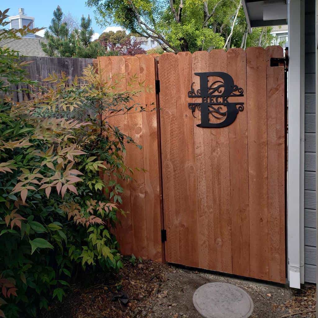 Wooden side yard gate with a decorative black metal monogram, surrounded by lush greenery and a gravel pathway