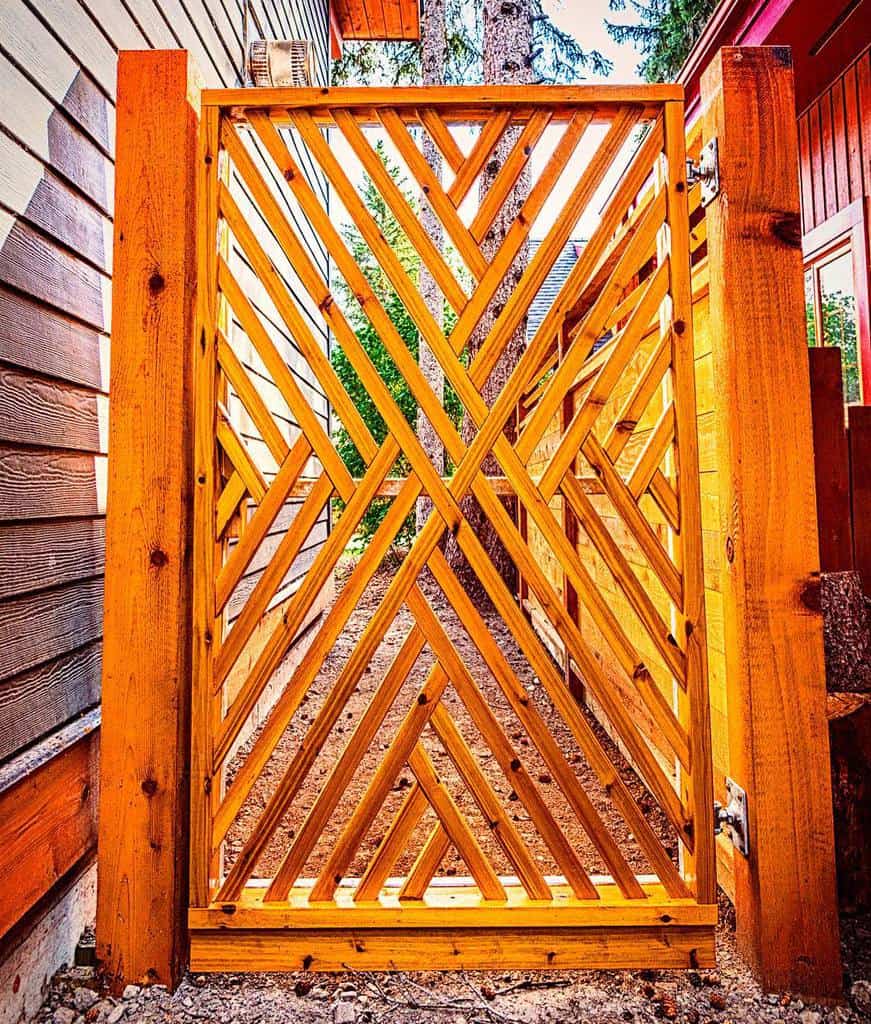 Wooden gate with diagonal slats and geometric pattern, framed by wood posts, leading to a tree-lined area