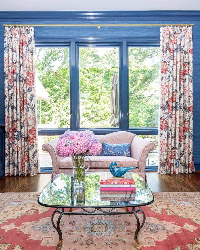 Glass coffee table with ornate metal legs, styled with books, a blue bird figurine, and pink hydrangeas, set in a vibrant living room with floral curtains