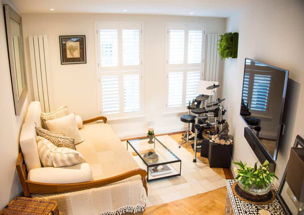 Bright living room with a sofa, glass coffee table, wall-mounted TV, and electronic drum set; white shutters on windows and a plant decor