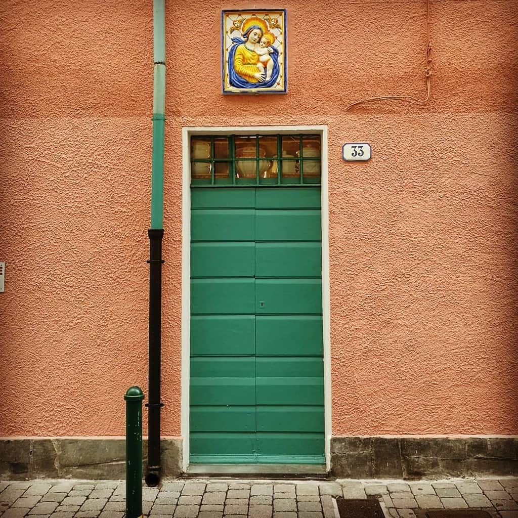 Green door on a peach-colored wall with a pipe beside it and a religious image of Mary and Jesus above, cobblestone path in front