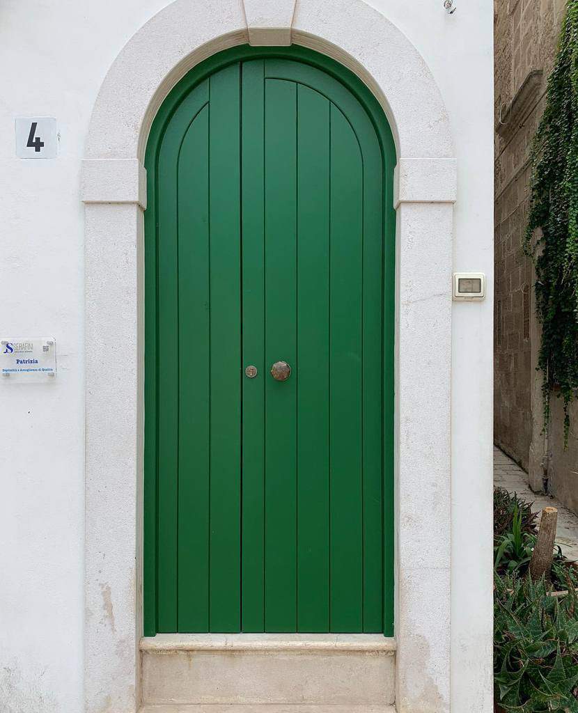 Arched shiplap green door with white stone frame set in a white wall, two circular handles at the center, house number 4 displayed nearby