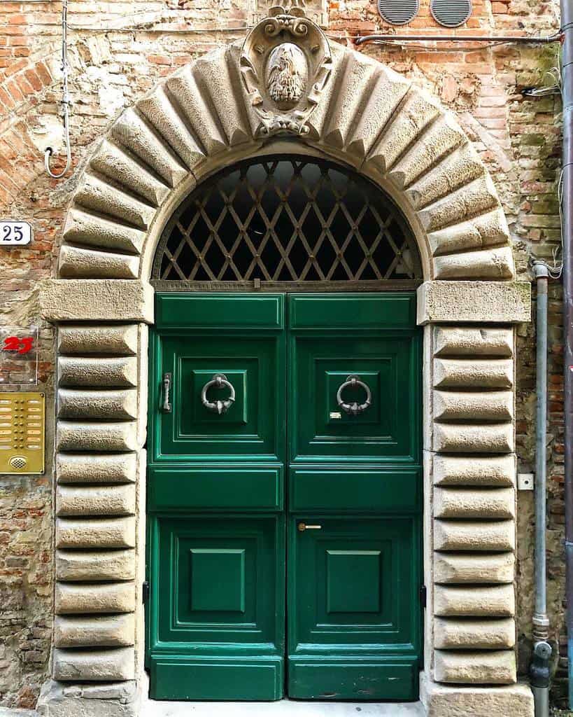 Ornate green double door with curved stone archway, decorative stonework, and window grate, set in an old brick wall