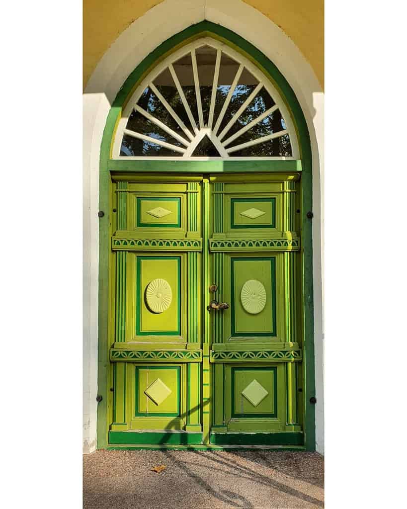 Ornate green door with decorative panels and a semicircular window above, set in an arched entryway with sunlight casting shadows