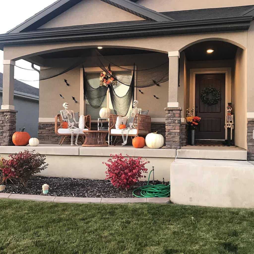 A porch decorated for Halloween with skeletons sitting on chairs, pumpkins, black netting, and a wreath, and a skeleton standing by the door