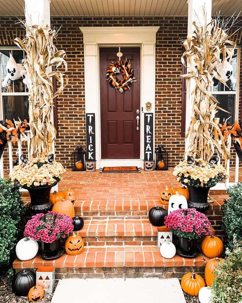 Festive Halloween porch with pumpkins, hay, mums, ghost decorations, and "Trick" and "Treat" signs flanking a maroon door