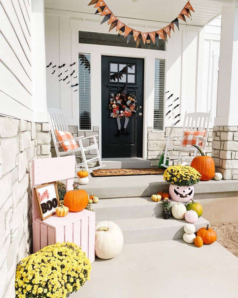 Front porch decorated for Halloween with pumpkins, yellow flowers, a "Boo" sign, and a black door wreath; two rocking chairs sit nearby