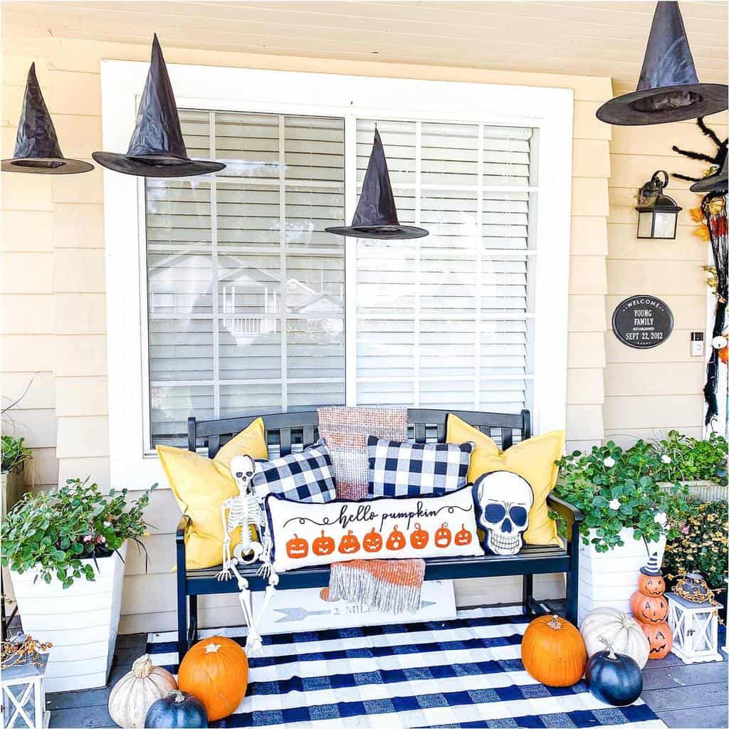 Front porch with Halloween decor: hanging witch hats, pumpkins, skeleton, plaid pillows, and a "hello pumpkin" sign on a black bench