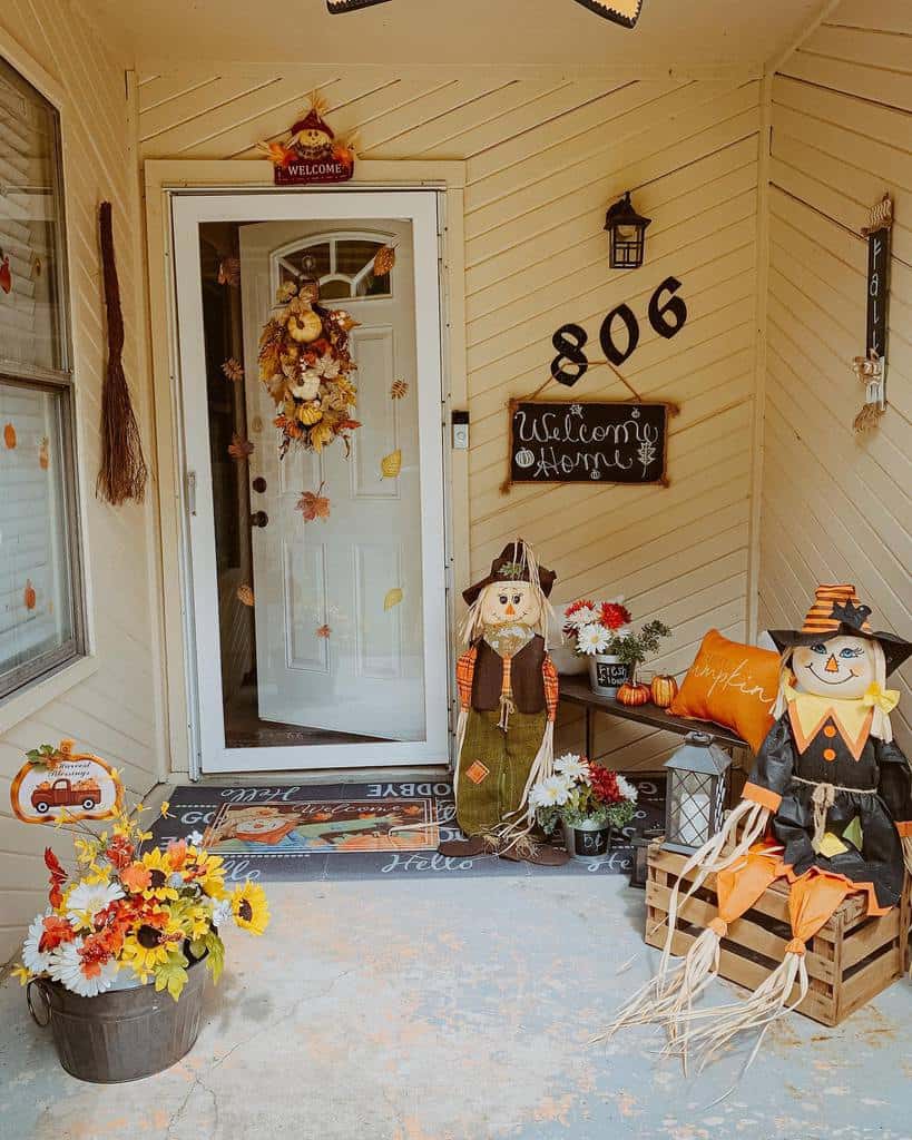 Festive autumn porch with scarecrows, flowers, pumpkins, and a "Welcome Home" sign next to a door adorned with a fall wreath