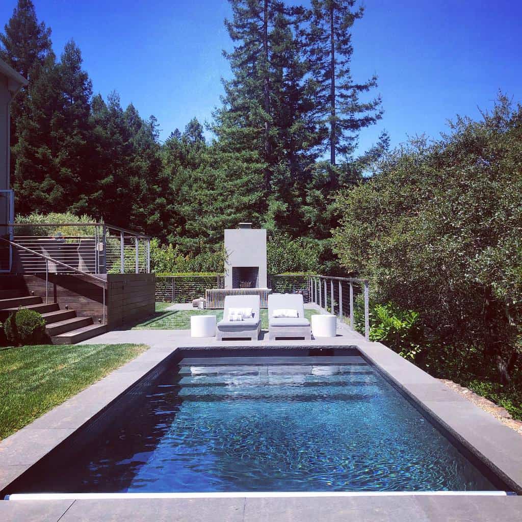 A rectangular outdoor pool with two white lounge chairs, surrounded by greenery and trees, under a clear blue sky