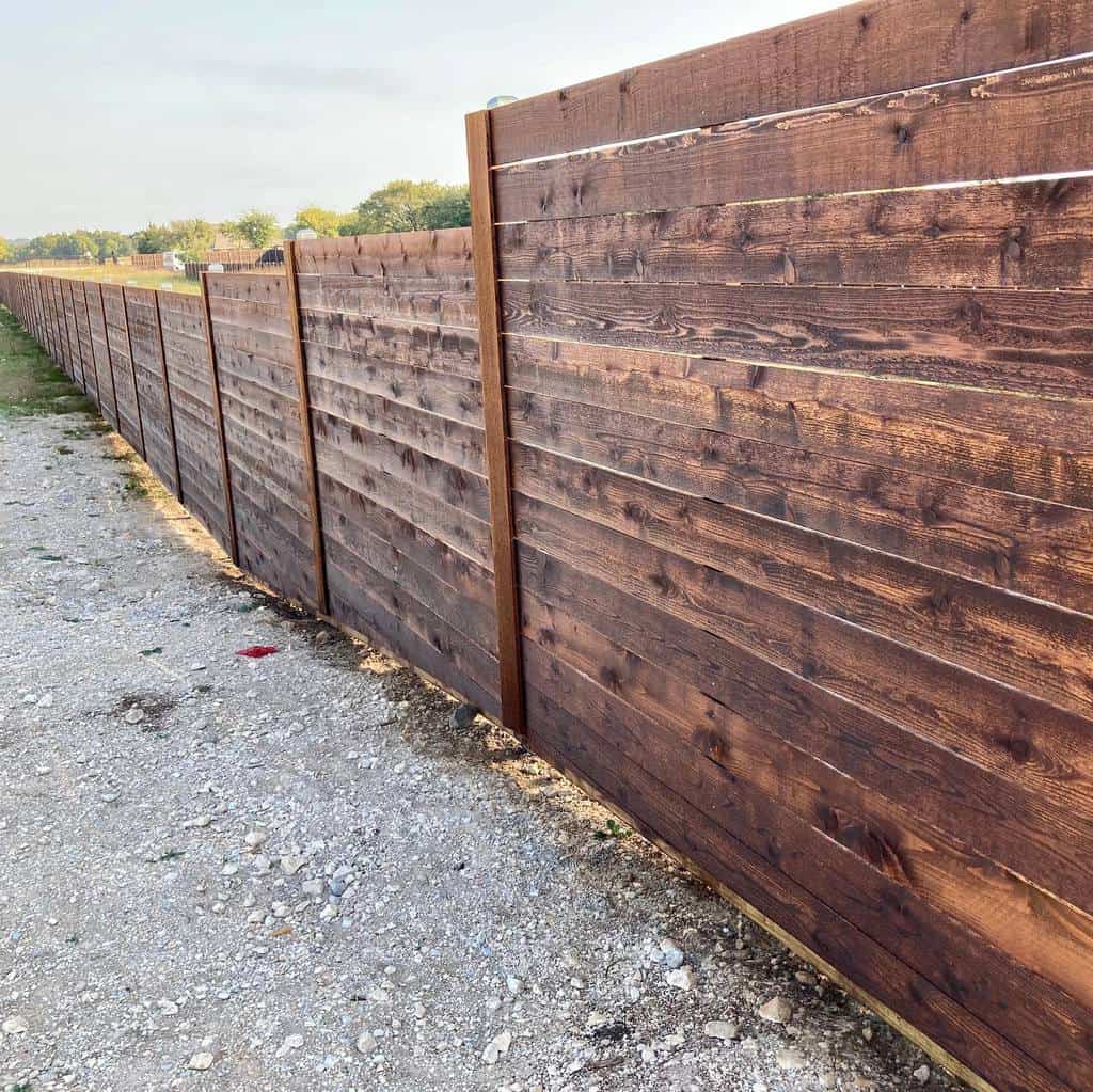 Long, dark-stained horizontal wooden fence providing privacy and a modern aesthetic, stretching along a gravel pathway