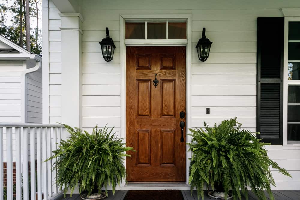 A wooden front door with a knocker, flanked by potted ferns on a porch, white siding, black lanterns, and nearby railings are visible