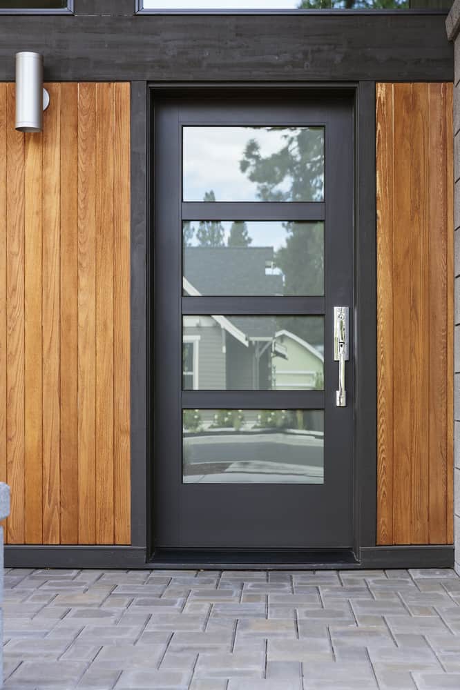 Modern front door with horizontal glass panels, surrounded by wooden siding and a metal frame, brick-paved entrance path