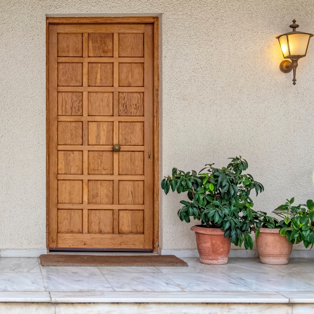 A wooden front door with a grid pattern, two potted plants on the right, and a lit wall lamp above on a stone porch