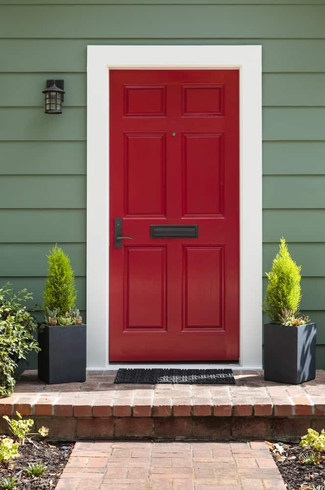 A bright red door with a white frame on a green house, flanked by potted plants on a brick pathway