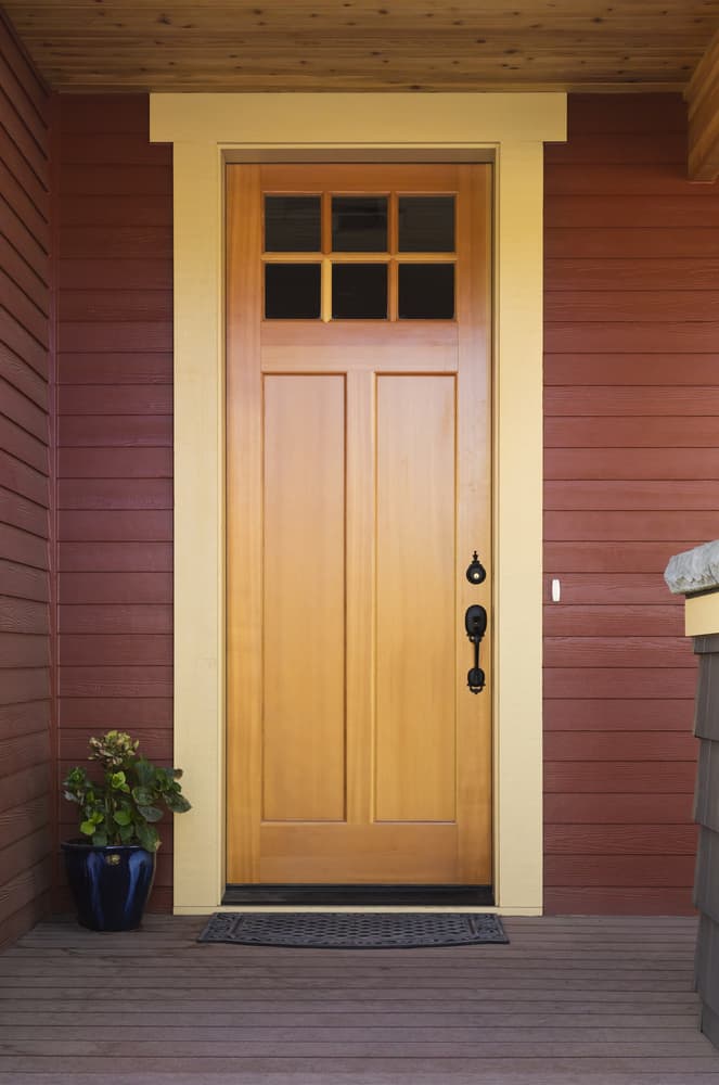 A wooden front door with square windows at the top, set in a red paneled wall, with a potted plant on the left and a porch floor mat