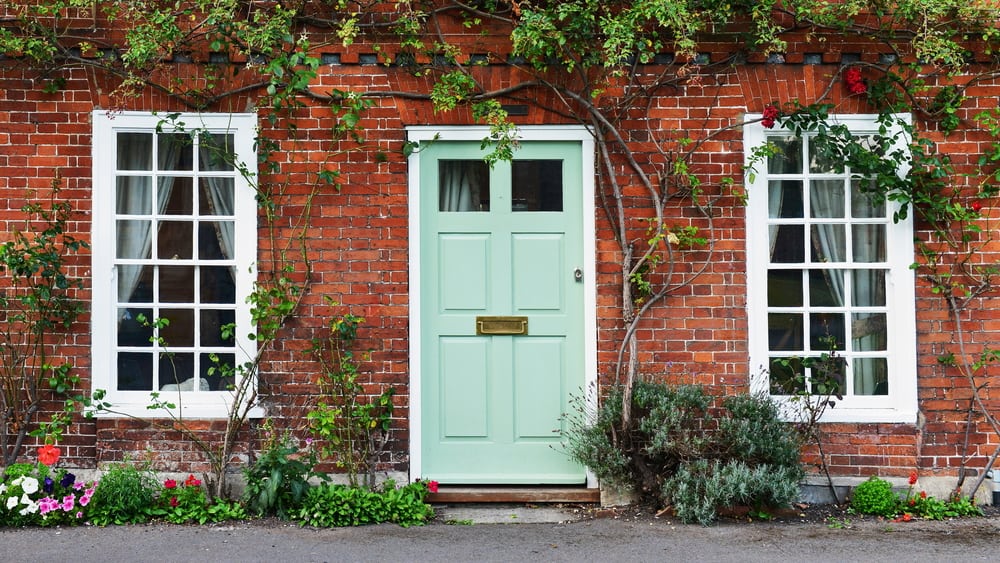 A charming brick house with a pastel green door, two large windows, and climbing plants surrounding the entrance