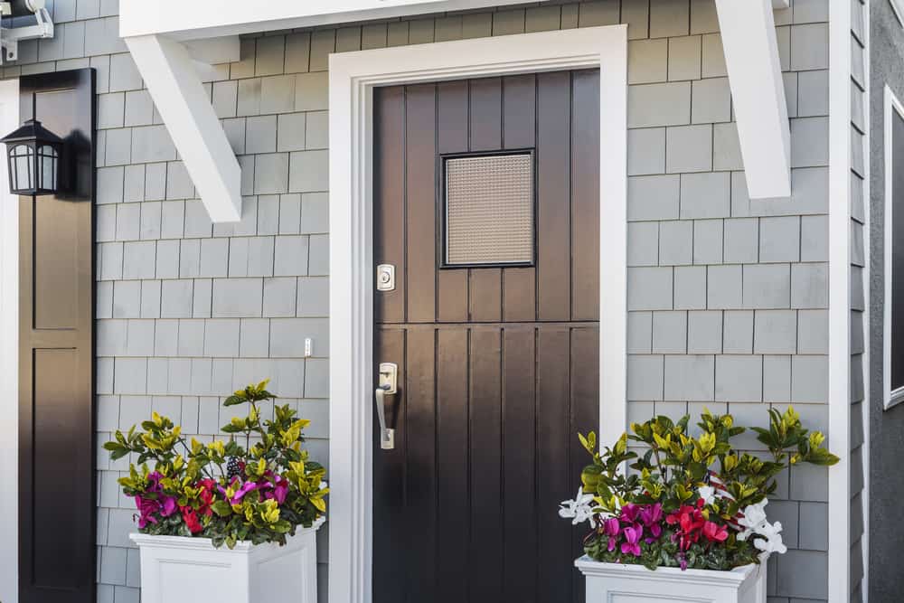 A black door with a small window, flanked by two planters filled with vibrant flowers, set against a gray shingled wall