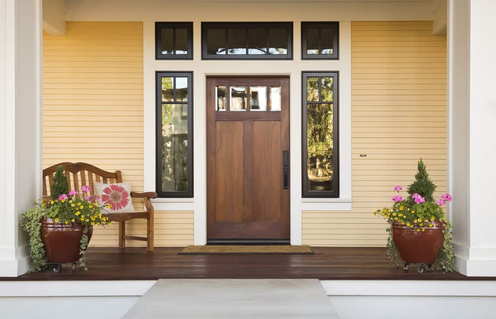 A wooden front door with a glass panel, flanked by plants in large pots and a wooden bench with a floral pillow, on a porch with yellow siding