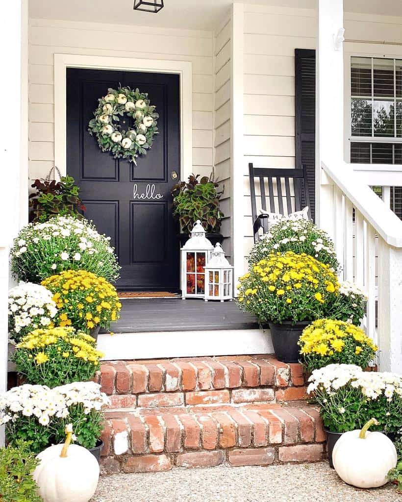 Front porch with a black door, wreath, "hello" sign, rocking chair, lanterns, and steps surrounded by white and yellow flowers and pumpkins