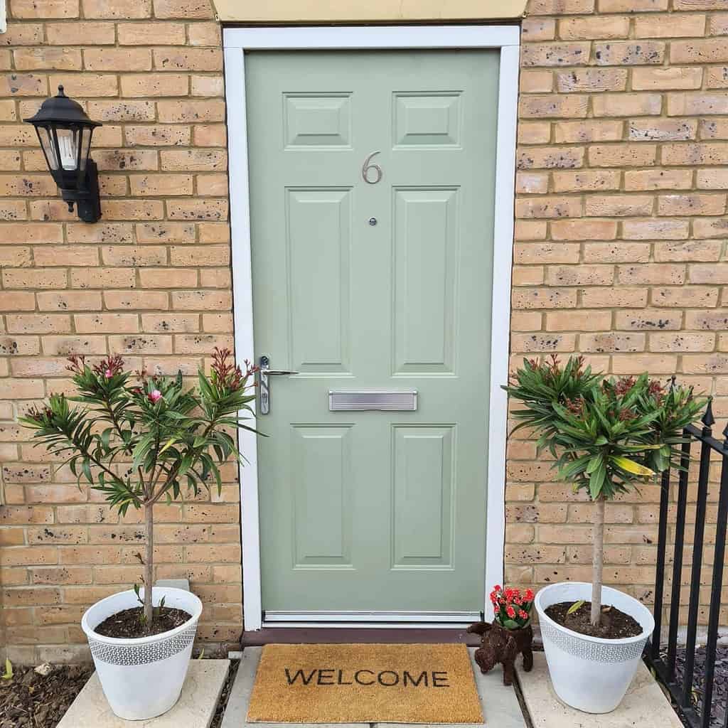 A light green door with a number 6, flanked by potted plants, a welcome mat, and a small decorative dog, against a brick wall