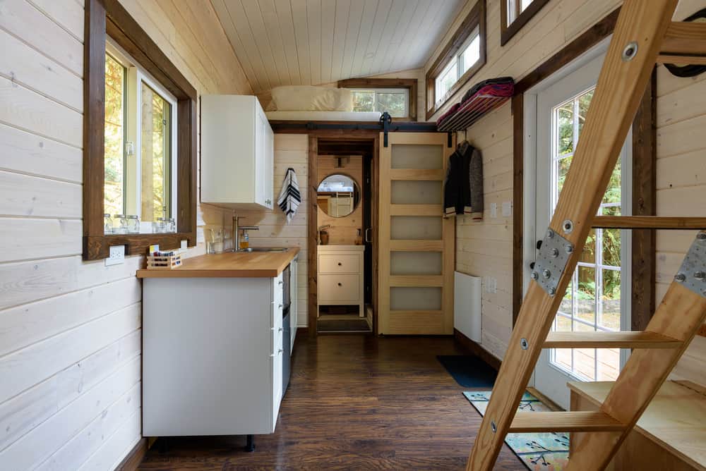 Bright tiny home interior with a lofted bed, sliding barn door, and compact kitchen. Large windows bring in natural light and warmth.