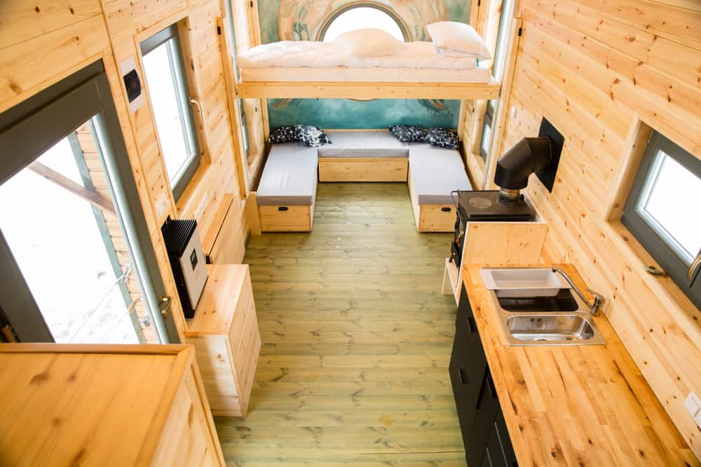 Warm wood-clad tiny home with a lofted bed, cozy seating, and a compact kitchen. A round window adds charm and natural light.