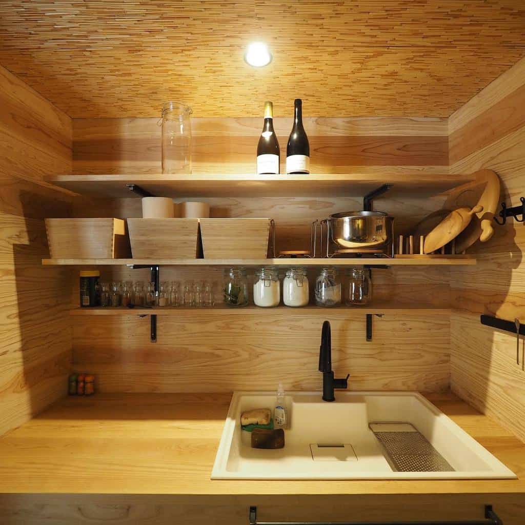 Warm wooden tiny house kitchen with open shelves, a deep sink, and black fixtures. Glass jars and baskets keep storage organized.