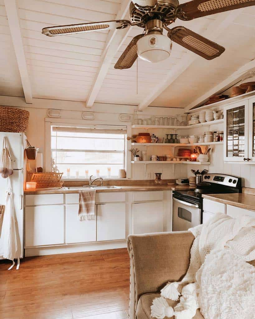 Cozy kitchen design for small spaces with white cabinets, wooden shelves, a ceiling fan, and soft lighting featuring warm tones and a beige armchair.