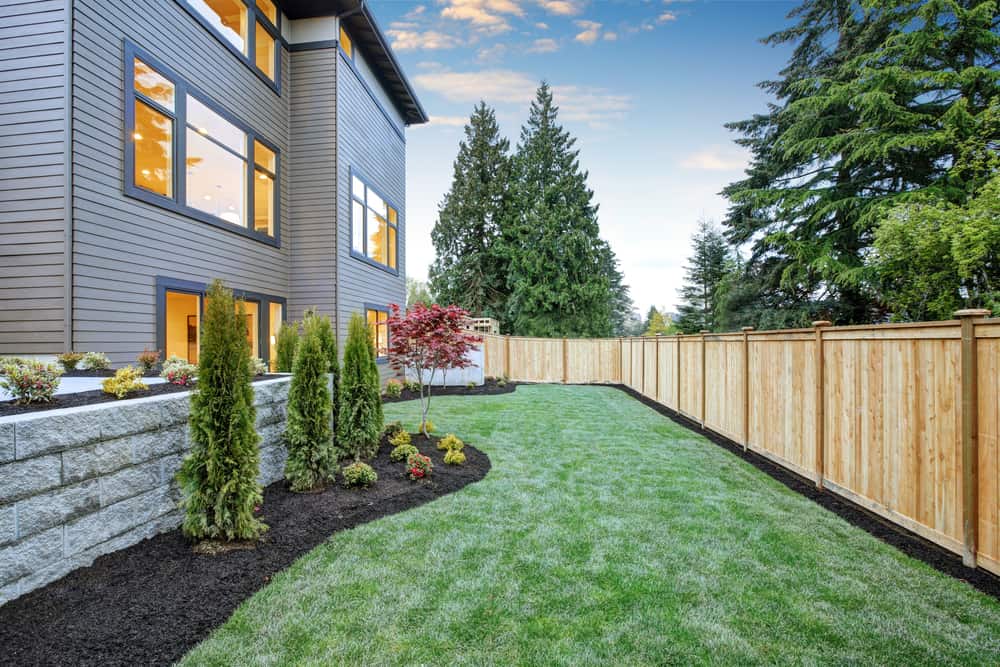 Modern house with large windows, lush green lawn, trees, and wooden fence under a blue sky