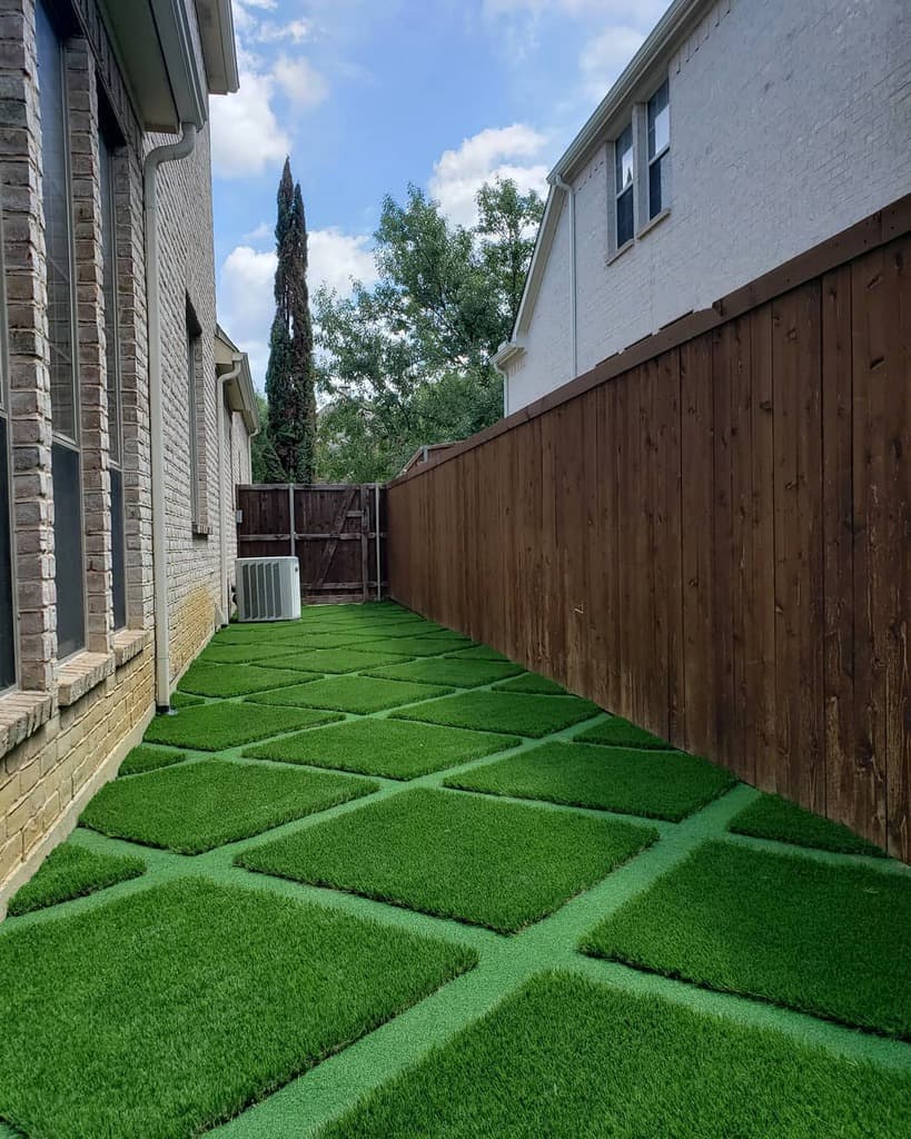 Side yard with square patches of artificial grass and concrete paths, next to a wooden fence and brick wall