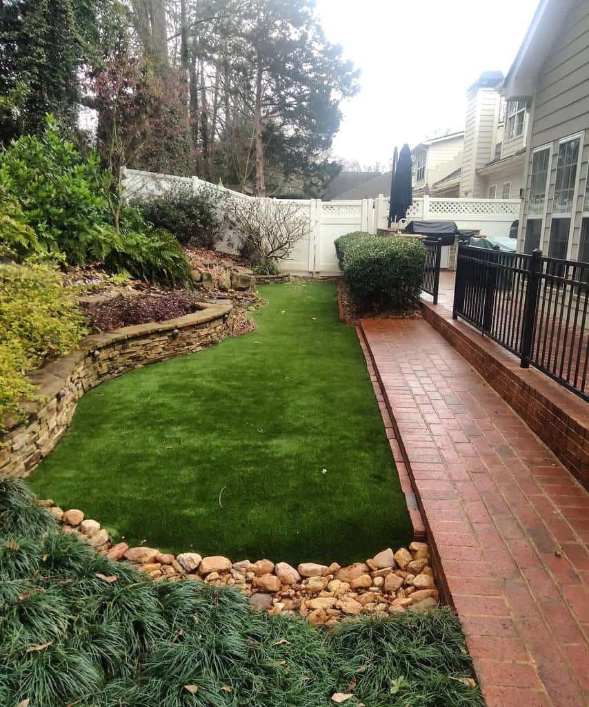 A small backyard with artificial grass, bordered by a stone wall and red brick walkway; enclosed by a white fence