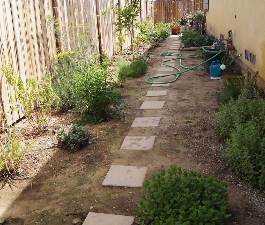 Narrow side yard with a dirt path, stepping stones, lush greenery, a wooden fence, and a garden hose near the house