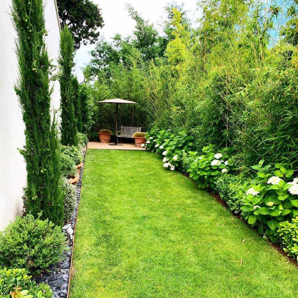 Narrow garden with lush green lawn, tall trees, white flowers, and a seating area with a bench and umbrella in the back