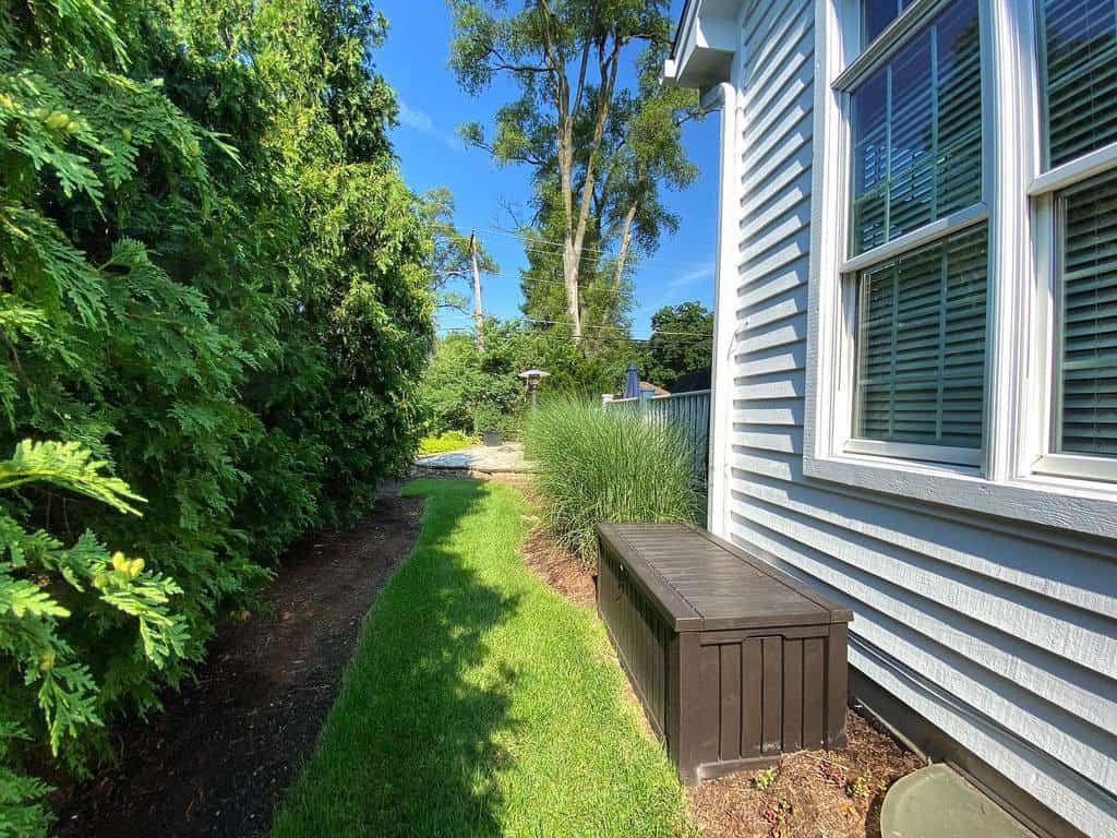 Side of a house with white siding, large windows, a brown storage bench, lush green grass, and tall trees