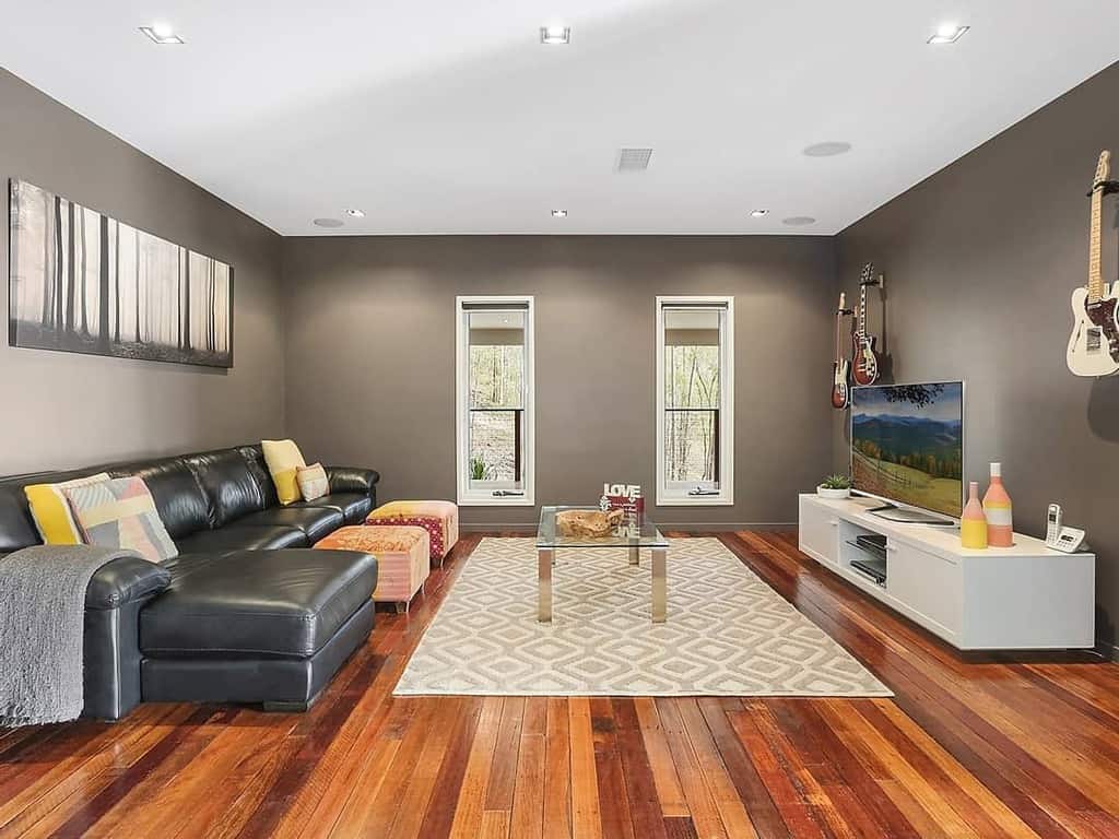 Modern living room with wall-mounted guitars, a black leather sectional, wooden flooring, a TV console, and a cozy, minimalist design