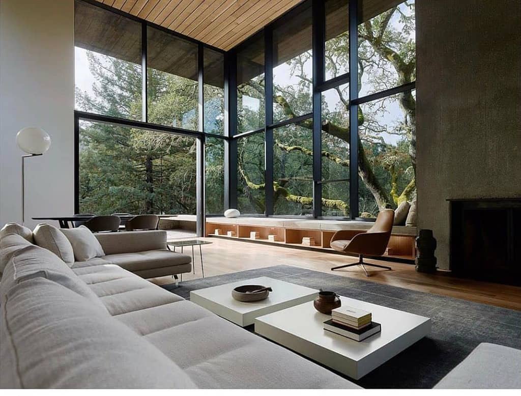 Modern living room with large windows, cream sofas, white coffee tables, and a view of trees. Minimalist and spacious design