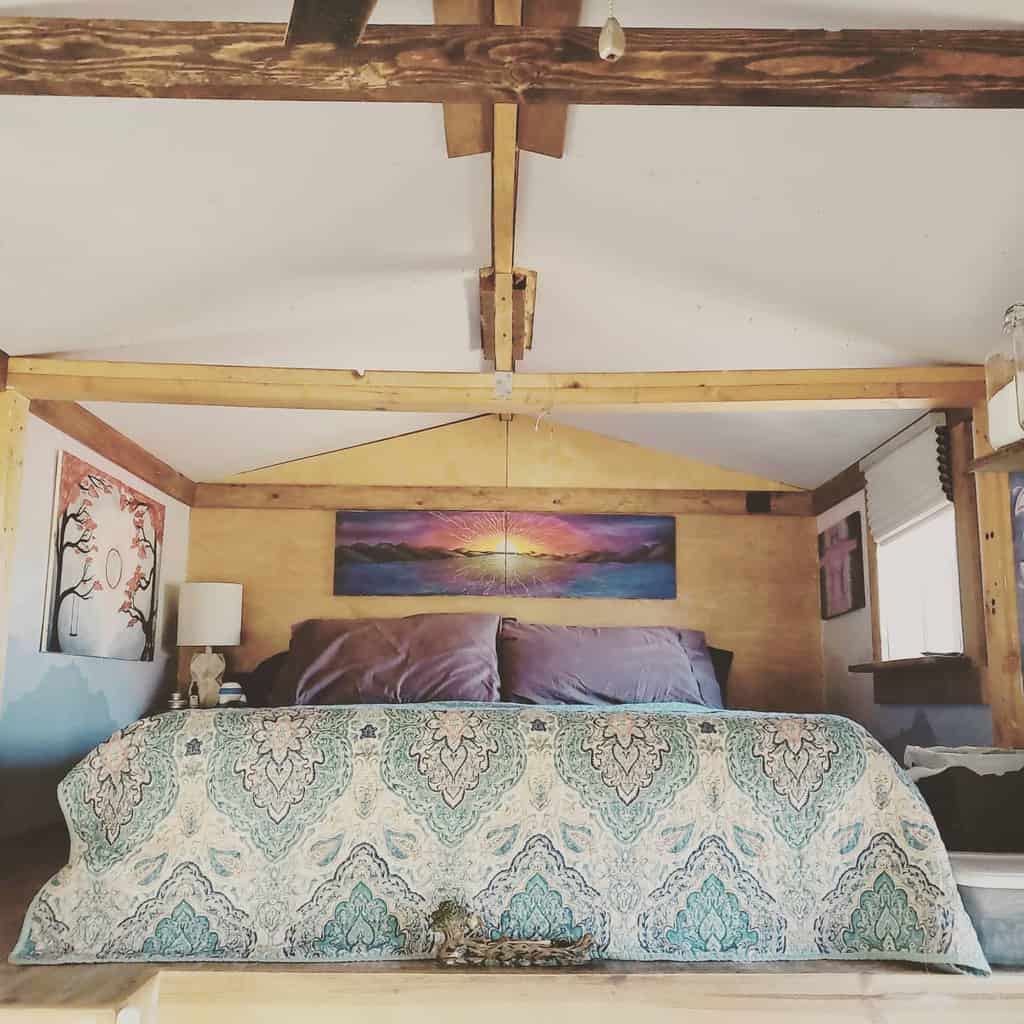 Rustic tiny house loft with exposed wooden beams, a patterned quilt, and warm lighting. Vibrant artwork adds character to the cozy space.