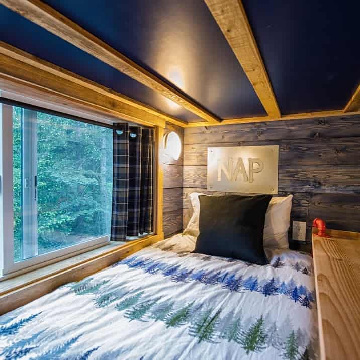 Cozy wooden cabin bed with "NAP" sign, forest bedding, and a large window with plaid curtain.