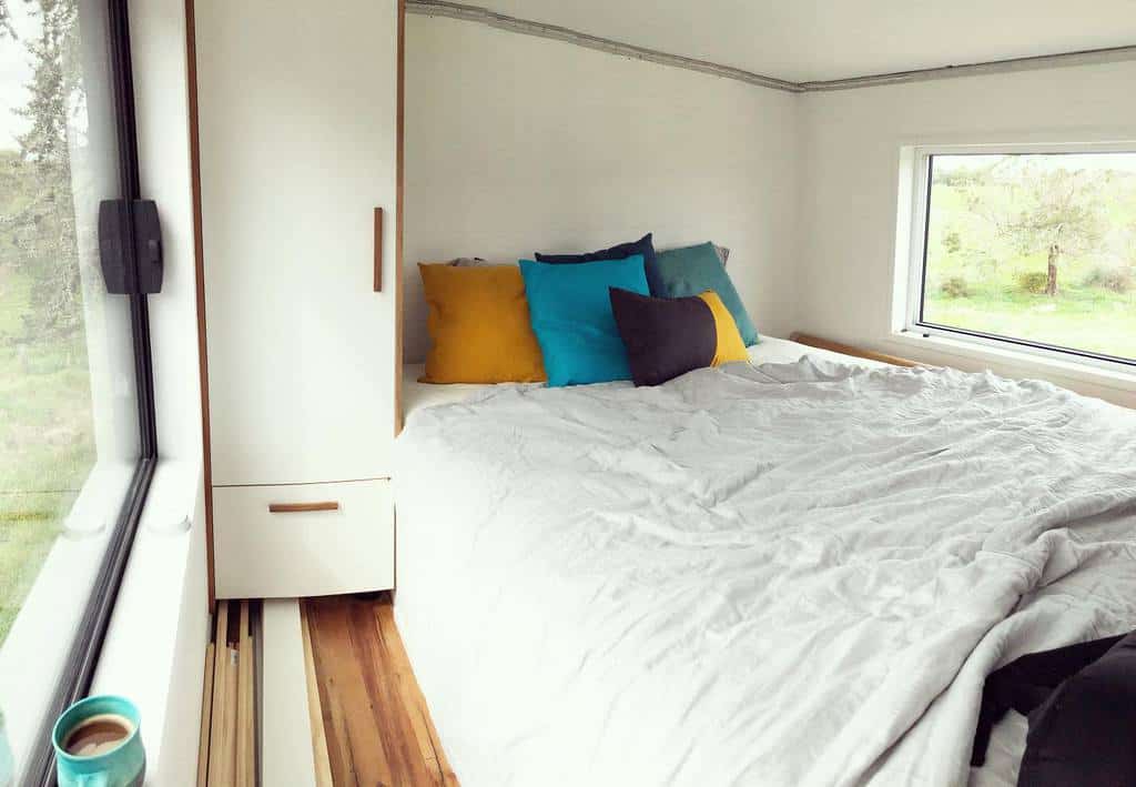 Cozy tiny house loft with large windows, a plush bed, and colorful pillows. A built-in wardrobe provides storage in the compact space.