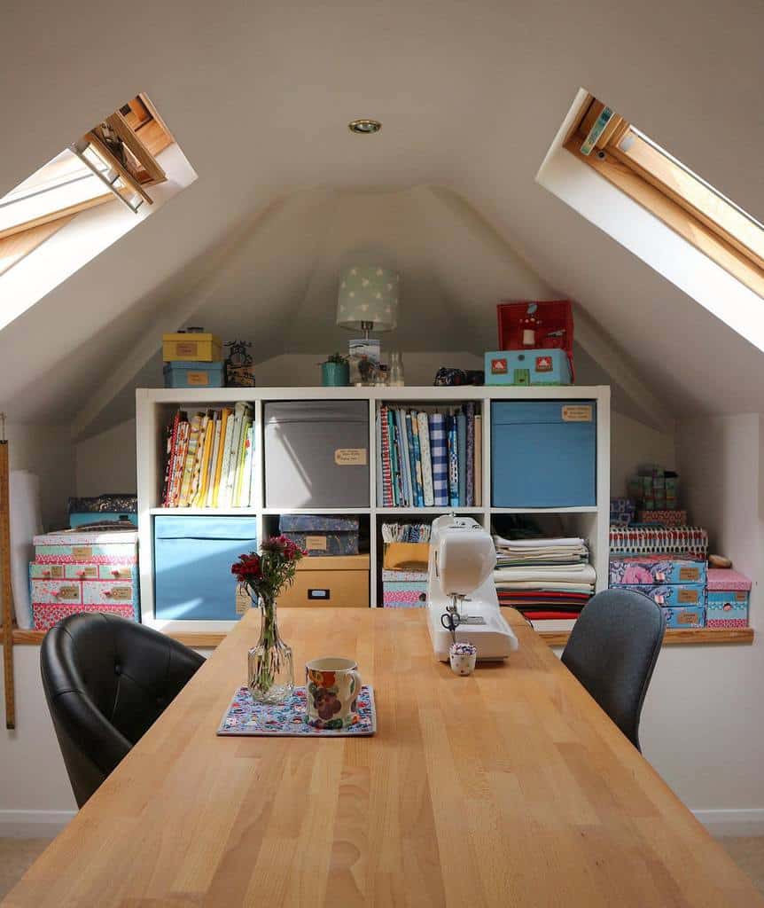 Cozy attic craft room with a wooden table, sewing machine, and shelves brimming with fabrics. Perfect small space design idea with slanted skylights.