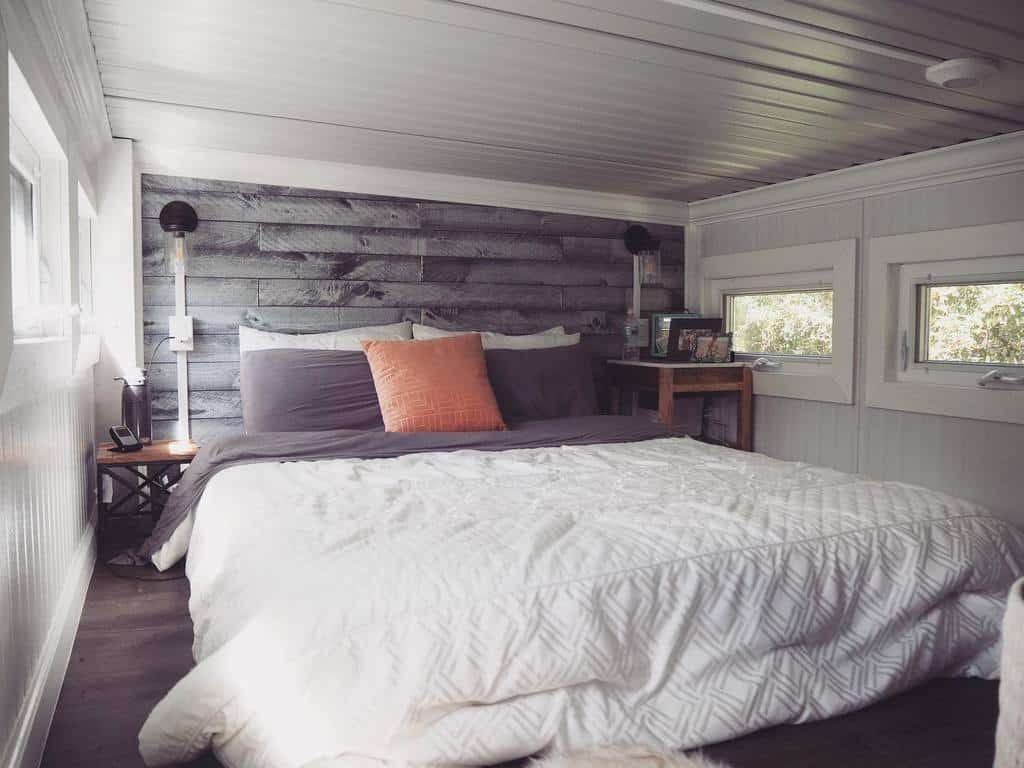 Cozy tiny house loft with a rustic wood accent wall, soft bedding, and warm lighting. Multiple windows bring in natural light.