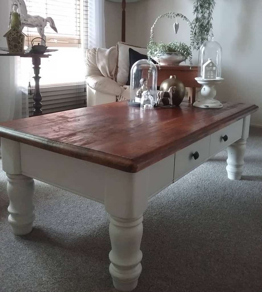 A rustic coffee table with wooden top and white legs, adorned with decorative cloches and ornaments, in a cozy living room