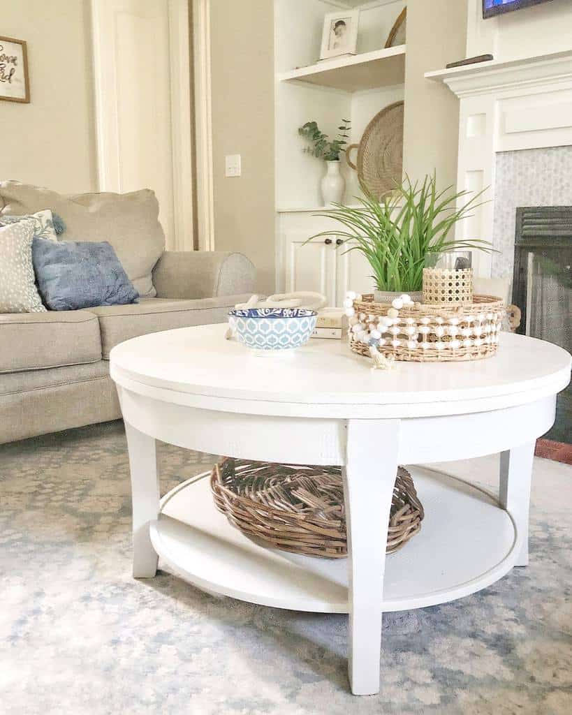 Round white coffee table with a lower shelf, styled with a woven tray, greenery, and decorative accents, set in a cozy neutral-toned living room