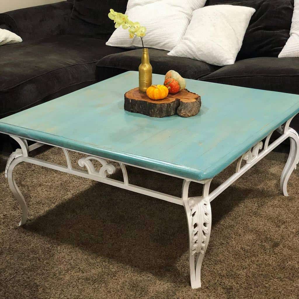 Square coffee table with a blue top and white ornate legs, decorated with a vase holding flowers and small pumpkins on a wooden tray