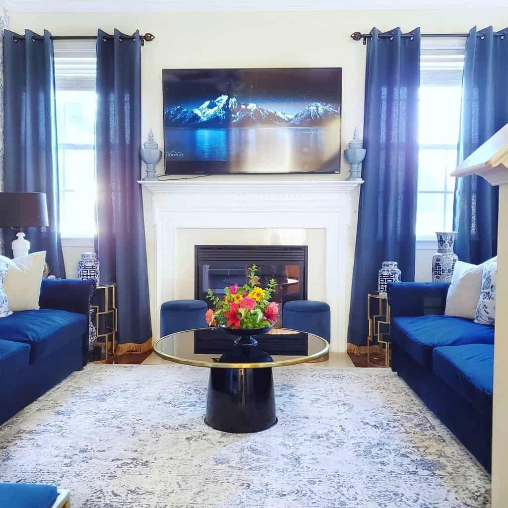 Stylish living room with blue sofas, a central black coffee table, ornate fireplace, flat-screen TV, and large windows with blue curtains