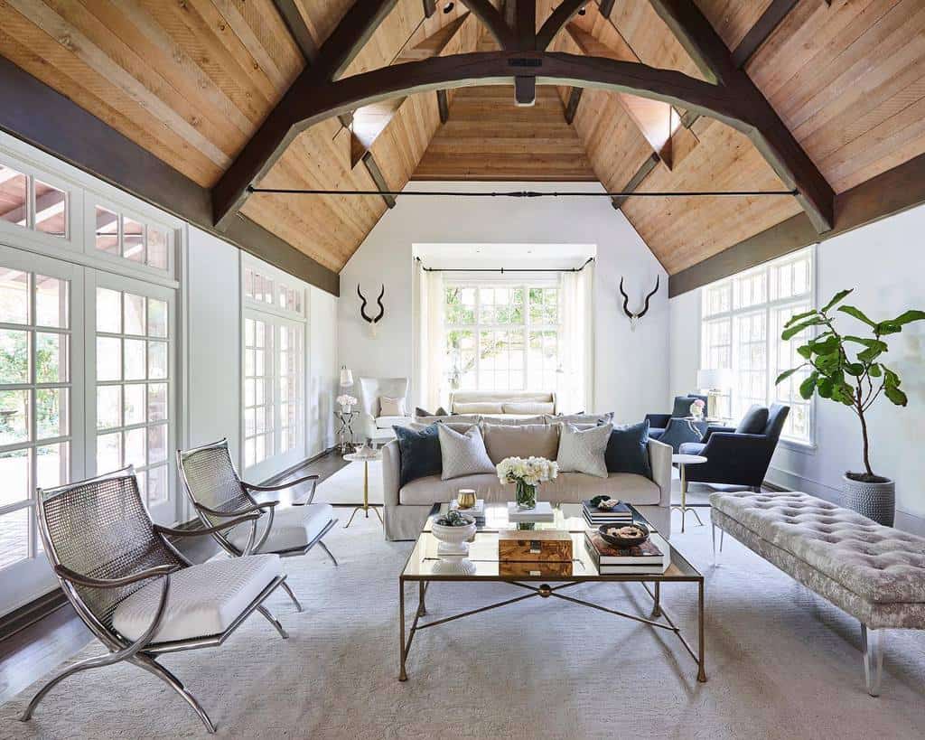 Elegant glass coffee table with a gold metal frame, styled with books, candles, and flowers, set in a spacious living room with vaulted wood ceilings