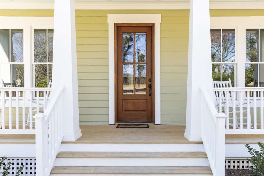 Front porch with a wooden door, white railing, and rocking chairs; yellow-green siding and a welcome mat on the steps